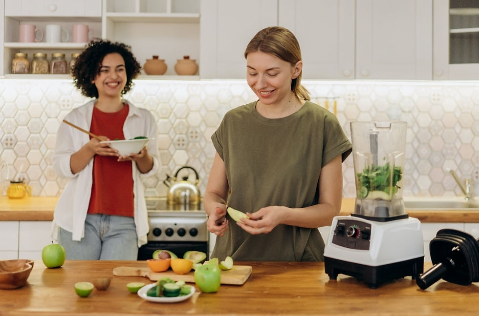 Two women in a kitchen; one peeling fruit beside a blender, the other eating salad and smiling. Light colors and a patterned backsplash.