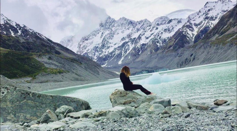 I Dream of Mangoes in black sitting by a frozen lake with snow capped mountains in the background