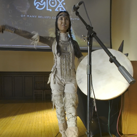 Snow Raven with a drum in one hand and a drum mallet in another outstretched hand