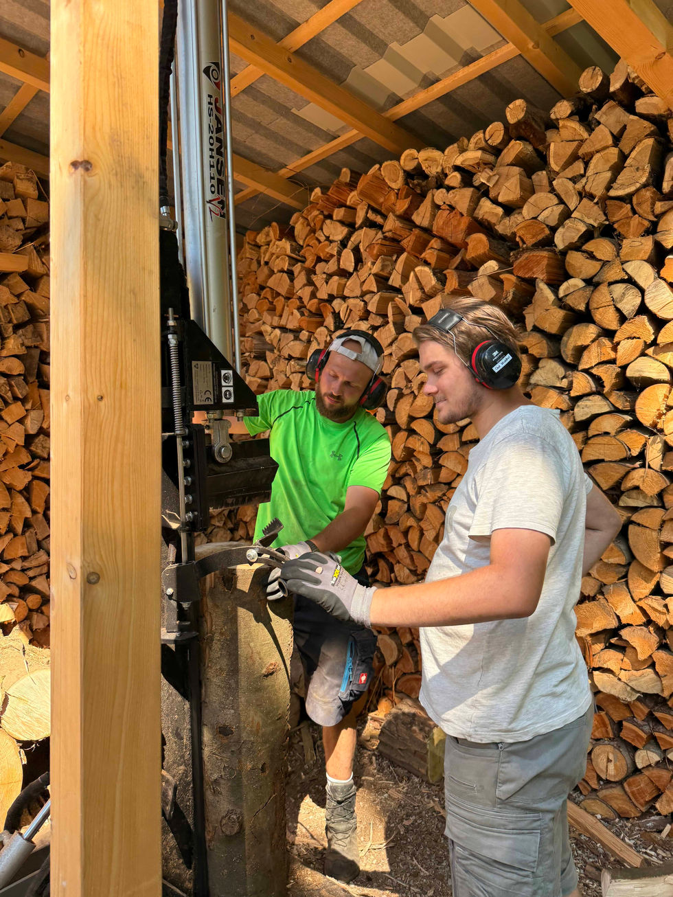 Helfer sägen, transportieren und spalten Holz im Wald, stapeln die Meterstücke ordentlich auf. Abends sitzen alle gemütlich beisammen und essen Flammkuchen am Feuer.