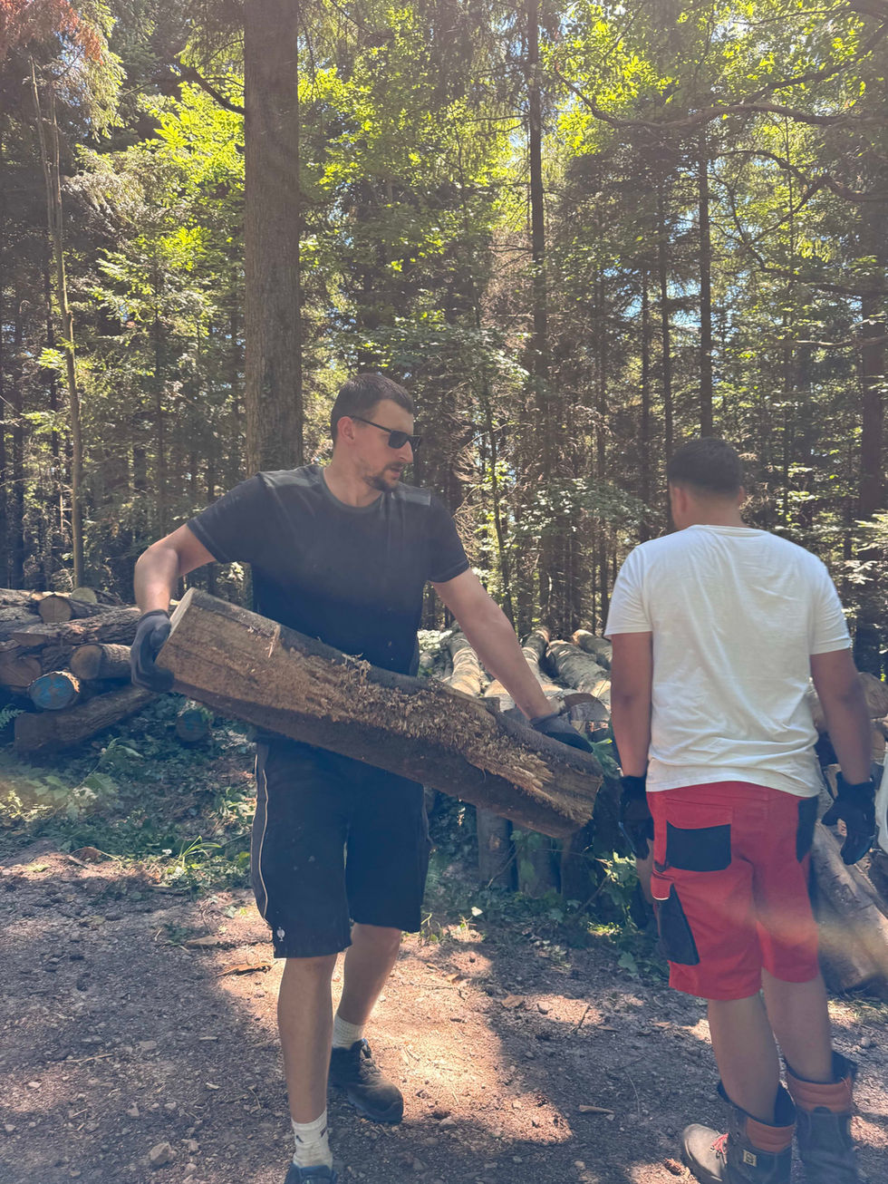 Helfer sägen, transportieren und spalten Holz im Wald, stapeln die Meterstücke ordentlich auf. Abends sitzen alle gemütlich beisammen und essen Flammkuchen am Feuer.
