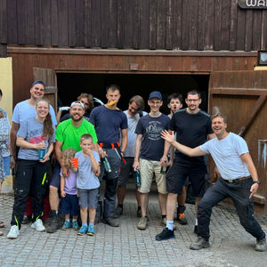 Helfer sägen, transportieren und spalten Holz im Wald, stapeln die Meterstücke ordentlich auf. Abends sitzen alle gemütlich beisammen und essen Flammkuchen am Feuer.