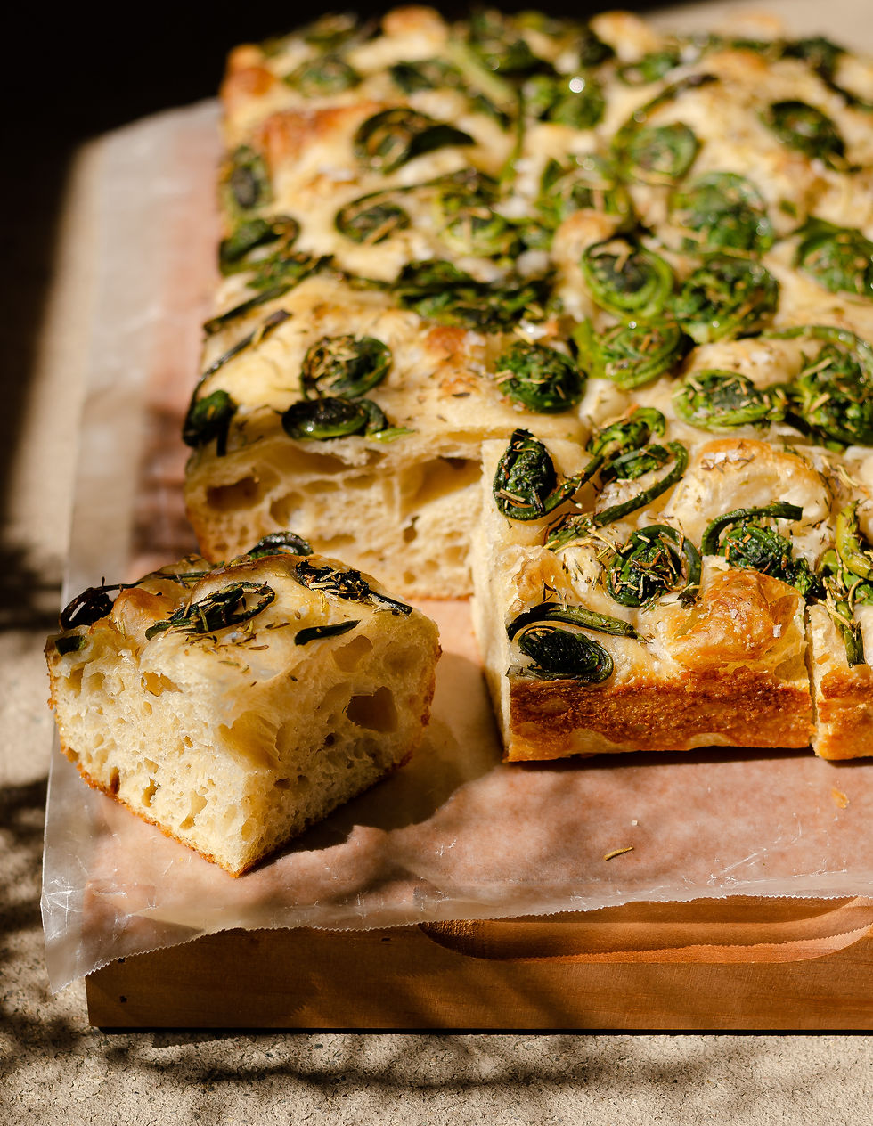 foraged fiddleheads and rosemary on top of sliced sourdough focaccia, placed on a wood board in the warm afternoon sun