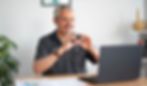 Man smiling, signing FAMILY into a webcam on a laptop. Indoor setting with plant, framed art, and shelves in the background.