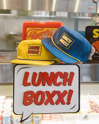 Yellow and blue corduroy hats with LUNCH BOXX patches displayed alongside retro lunchbox.