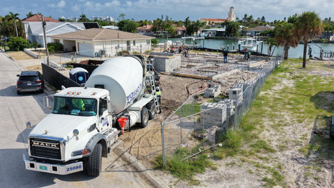 Aerial view of a waterfront home under construction, with workers on a concrete foundation beside docks, boats, and palm-lined houses.