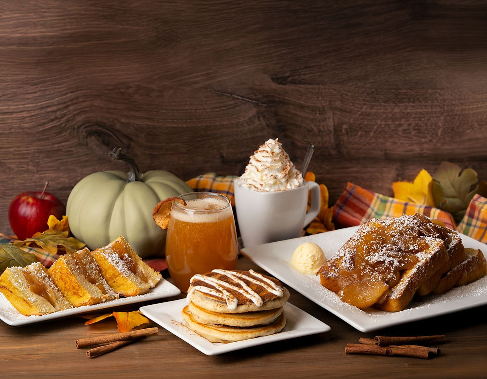 Assortment of Keke's fall dishes on a rustic wood table, including caramel apple French toast, a stack of cinnamon roll pancakes, a whipped-cream-topped pumpkin drink, and an apple cider mimosa, surrounded by mini pumpkins, apples, and fall leaves