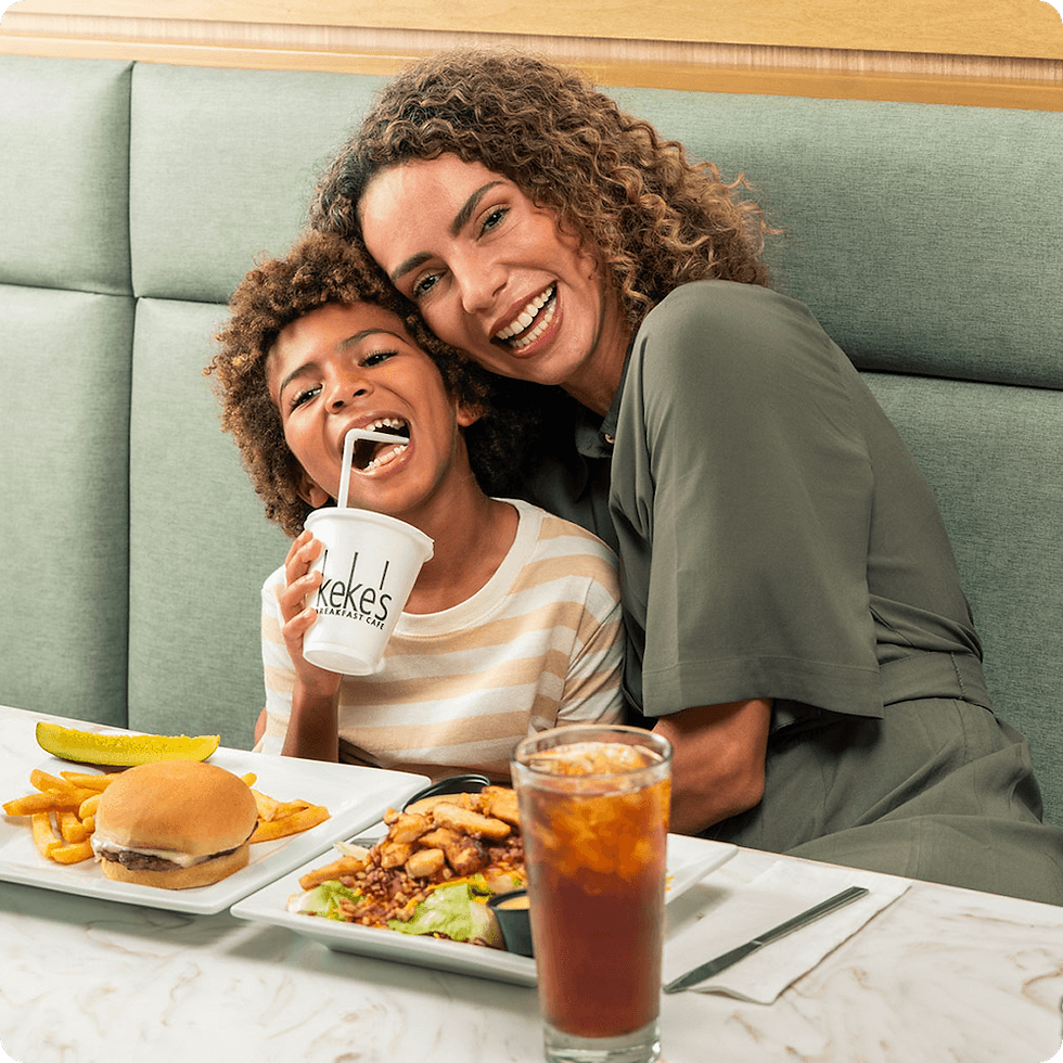 Mother and child smiling in a Keke's Breakfast Cafe booth with a kids cheeseburger, fries, and Keke's branded drink cup