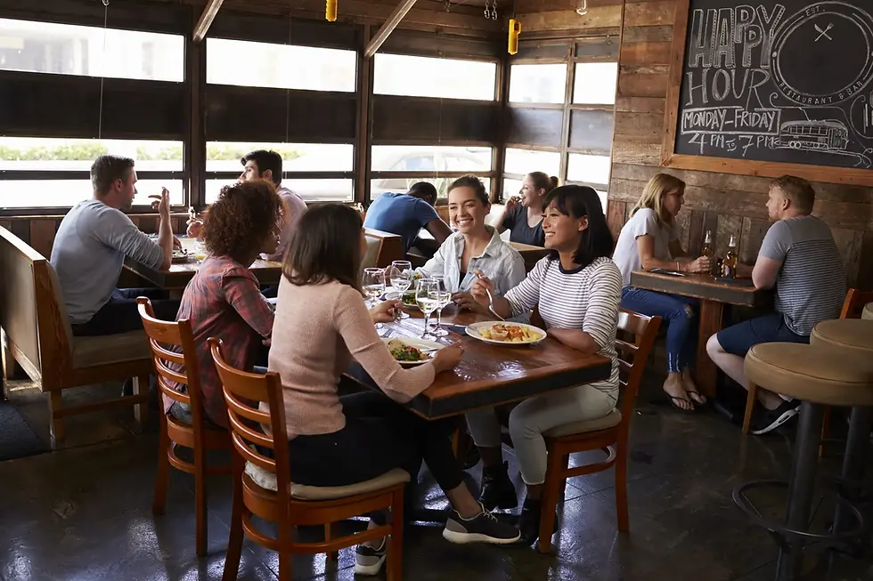 Friends enjoying a meal at a rustic restaurant. Lively conversation, wine glasses on tables, "Happy Hour" sign on chalkboard in the background.
