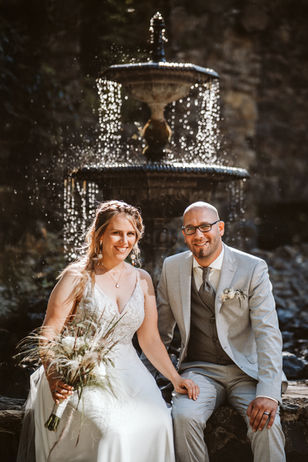 Romantisches Hochzeitsfoto vor einem historischen, plätschernden Brunnen, dessen Wasser im Gegenlicht glänzt. Das Brautpaar sitzt lächelnd auf einem Stein, die Braut hält einen rustikalen Hochzeitsstrauß.