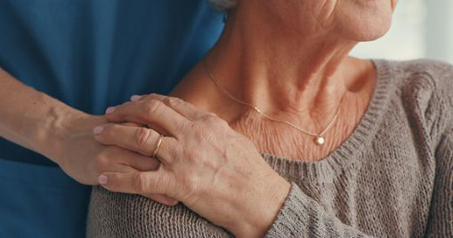 A younger woman holds the hand of an older woman who is seated.  This image illustrates the connection and support of an adult child caregiving a person living with dementia.