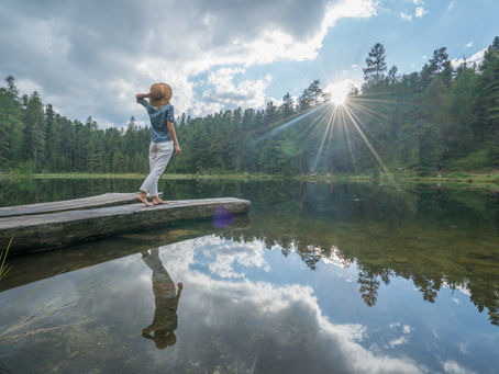 A woman stretches on a dock over a calm lake while watching the sun set behind a forest of trees