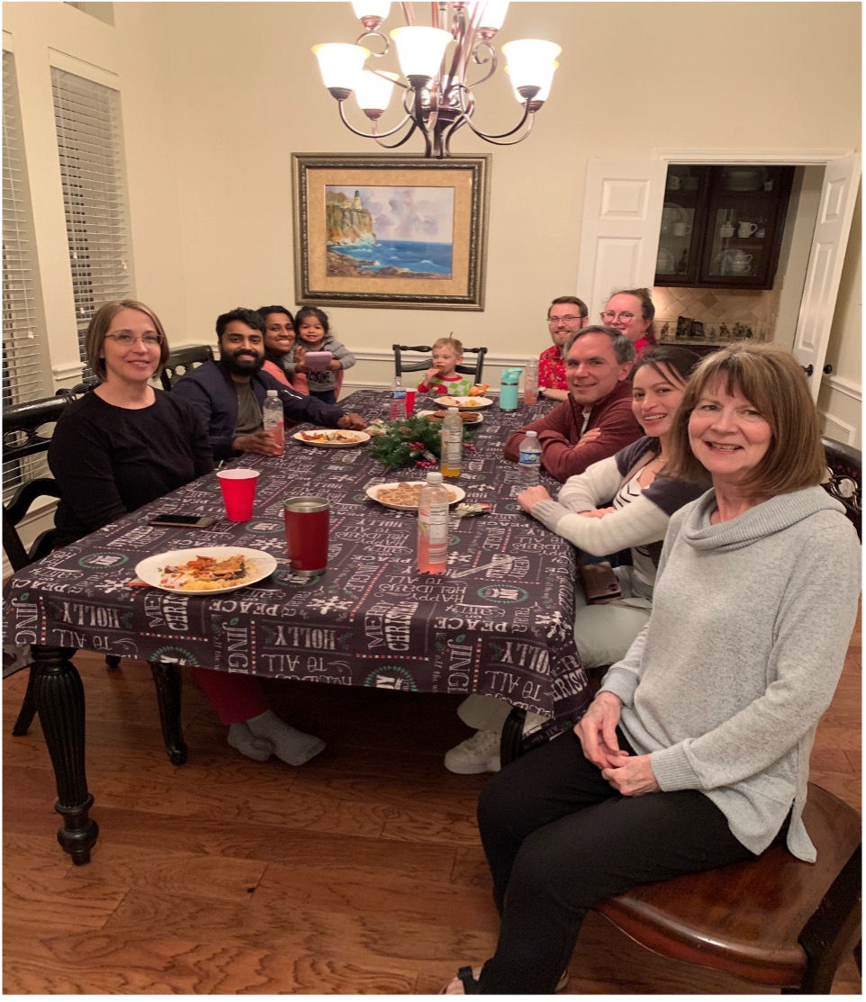 A group of people smiling around a dining table with food, under a chandelier. Festive tablecloth, wood floor, and framed art in background.