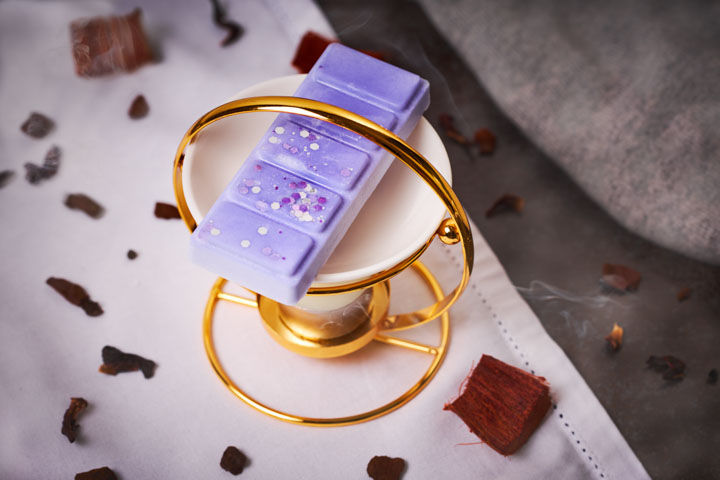 Purple wax melt on white and gold burner, on white tablecloth, with burned smoky wood and cashmere.