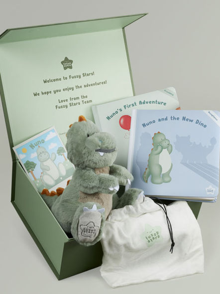Green dinosaur soft toy positioned in the front left corner of a box facing right, showing branding on toy animal paw, with two padded covers kids book to the side, a soft cotton bag in front and a card at the back.