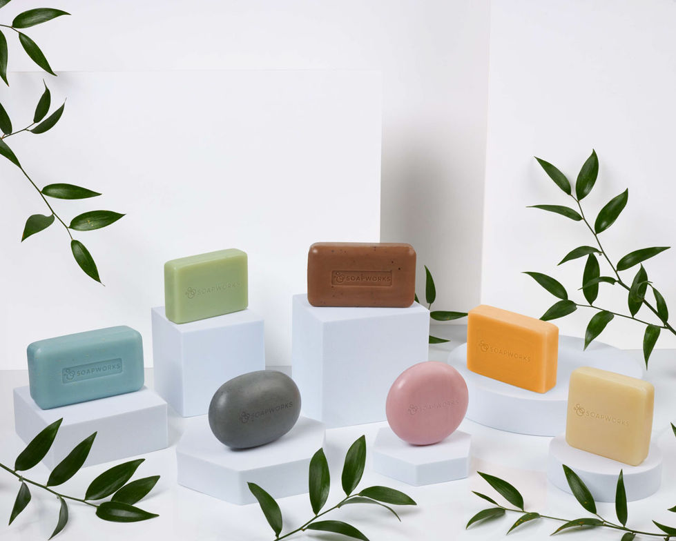 Soapworks Off The Shelf range soap body and shampoo bars, on white plinths and white background, surrounded by leaves.