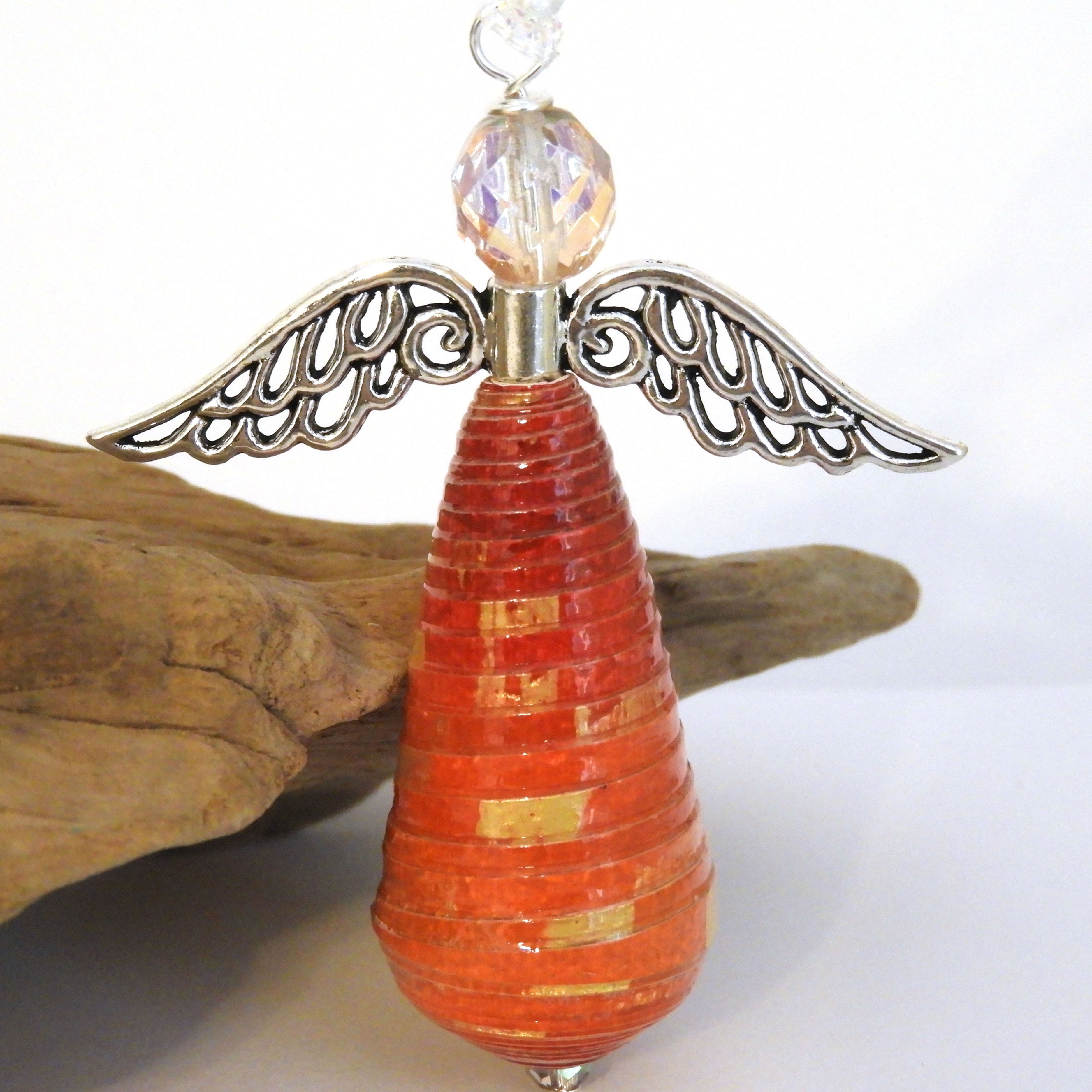 Red, orange, and gold ombre paper bead angel ornament standing in front of a piece of driftwood.