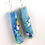 Thumbnail: Blue, white, and gold abstractly painted paper bead earrings hanging on a white background.