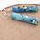 Thumbnail: Blue, white, and gold abstractly painted paper bead earrings on a wood background.