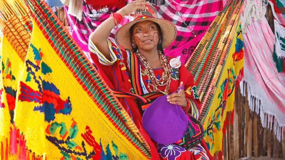 A woman in vibrant traditional attire sits in a colorful hammock market, smiling. She holds a purple yarn ball, surrounded by woven textiles. INDIGENA GUAJIRA