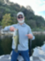 Man in sunglasses and cap on a boat holds a fish, pointing at it. He's wearing a gray shirt; forested shoreline in background.