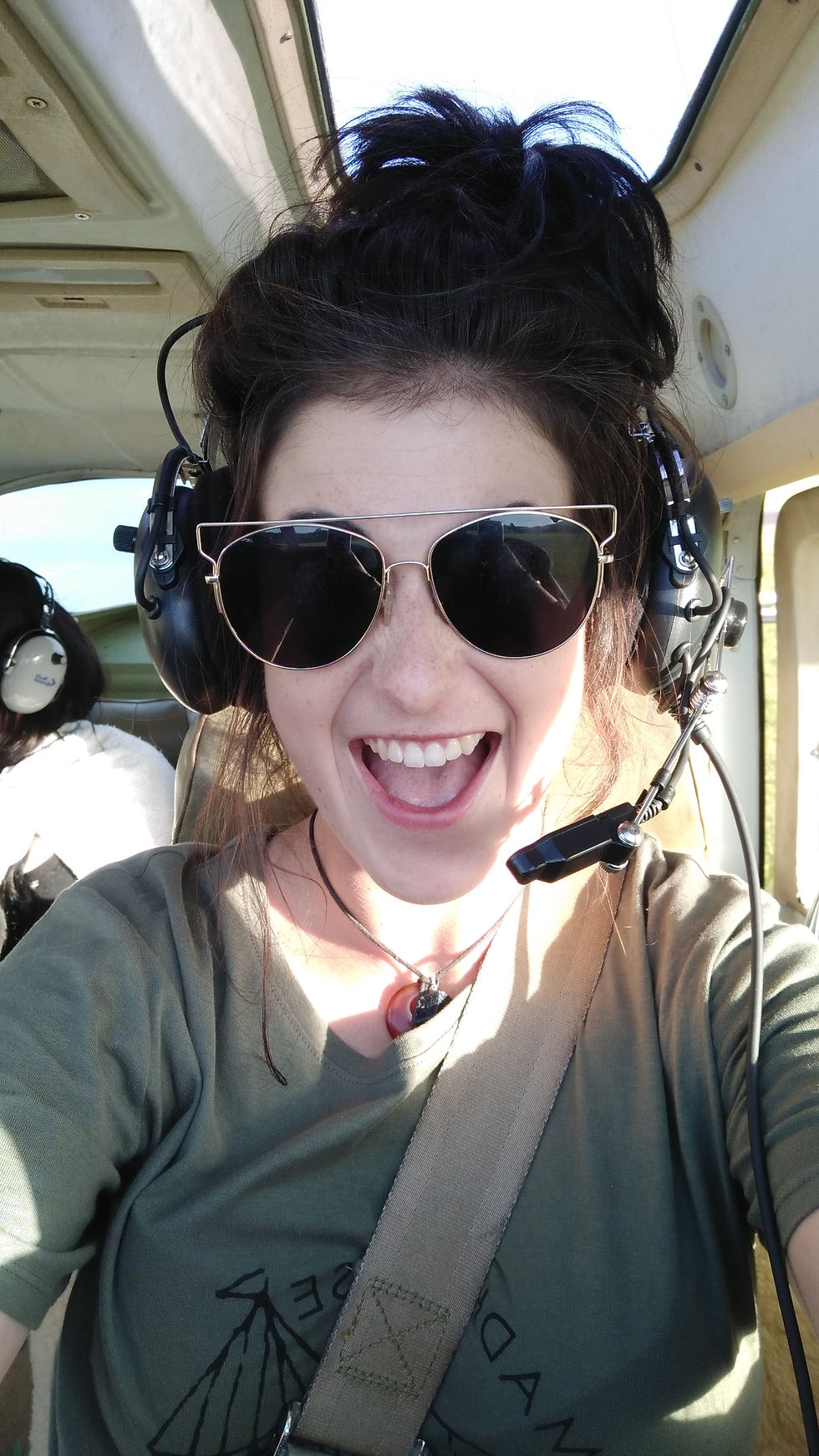 Front on selfie photo in the cockpit of Jess. She is wearing khaki green t shirt, aviator sunglasses, black hair tied up in a bun, with the headphones and with a microphone coming out towards her mouth. She has her mouth wide open with excitement.