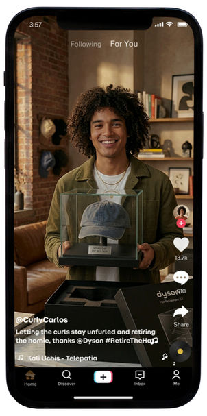 A digital mockup of a smartphone displaying a TikTok video from the account @CurlyCarlos. The video features a smiling man with curly hair wearing an olive green shirt, holding a glass display case that contains a faded blue baseball cap. The Dyson "Hat Retirement Kit" box sits open in the foreground. The on-screen caption reads, "Letting the curls stay unfurled and retiring the homie, thanks @Dyson #RetireTheHat," with the song "Telepatía" by Kali Uchis as the background audio. The standard TikTok interface elements—including the like, comment, and share icons—are visible on the right side.