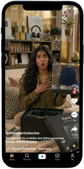 A digital mockup of a smartphone displaying a TikTok video from the account @PriyaHairCollective. In the video, a woman with long, wavy dark hair looks surprised, holding one hand to her chest while showcasing a navy blue beanie encased in a glass display box. In front of her is the open Dyson "Hat Retirement Kit" packaging. The caption on the screen reads, "She served for a while, but @Dyson serves harder. #RetireTheHat," with the song "Espresso" by Sabrina Carpenter listed as the background audio. The TikTok interface includes familiar icons for likes, comments, and sharing on the right side of the screen.