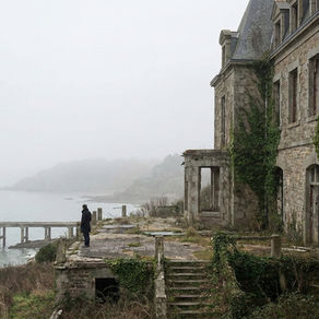 A solitary figure stands on a misty coastal cliff, gazing toward the sea next to a weathered, ivy-clad stone building. The scene evokes a sense of nostalgia and mystery, with the fog shrouding the landscape in a tranquil, ethereal ambiance.