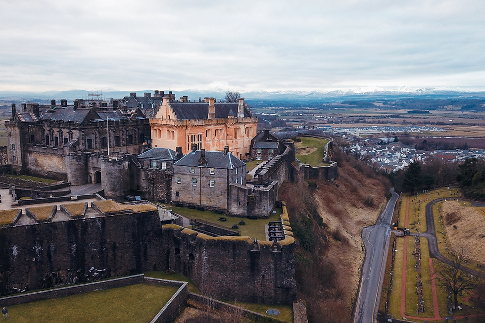 A historic castle on a hill overlooks a town and snowy mountains under a cloudy sky.