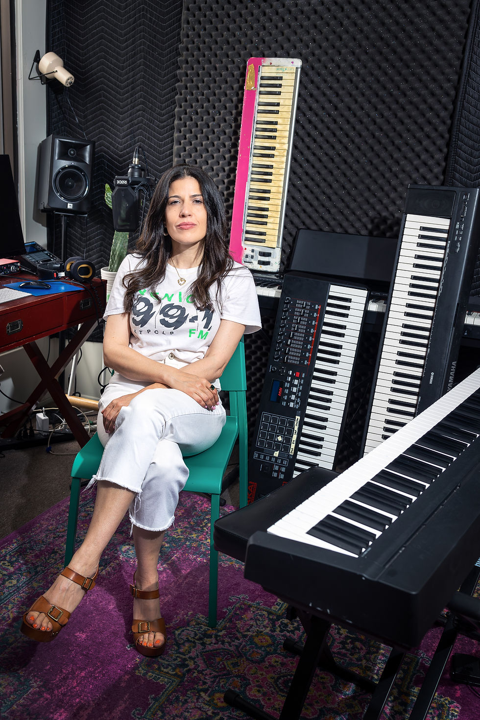 Woman sits on a green chair in a music studio with keyboards. Foam panels line the walls. She's wearing a white shirt and jeans.