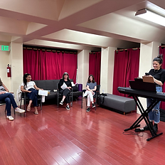 Group Singing Class in a large classrooms in Los Angeles with red curtains, 4 singers sitting in chairs and vocal coach standing at digital piano.