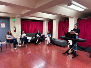 Singing for Fun: In-Person Singing Classes for Adults in Los Angeles, CA. This pictures is a voice teacher at a piano teaching a group of students sitting in a class room.