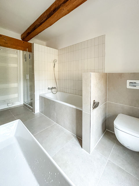 Modernes Badezimmer mit beiger Boden- und Wandfliese. Links eine Badewanne mit Handbrause, daneben eine verglaste Dusche. Rechts ein halbhoher Fliesen­trenn­wand und ein wandhängendes WC. Sichtbare Holzbalken an der Decke.