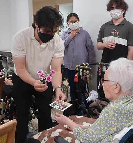 老人ホームを訪問したJICEFメンバーが、花と手作りのカードを手に、高齢の入居者に温かい交流の時間を提供している様子