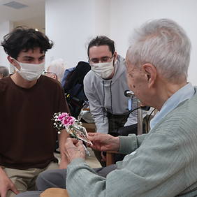 介護施設で、学生ボランティアが入所者に花を手渡しながら笑顔で交流している様子。