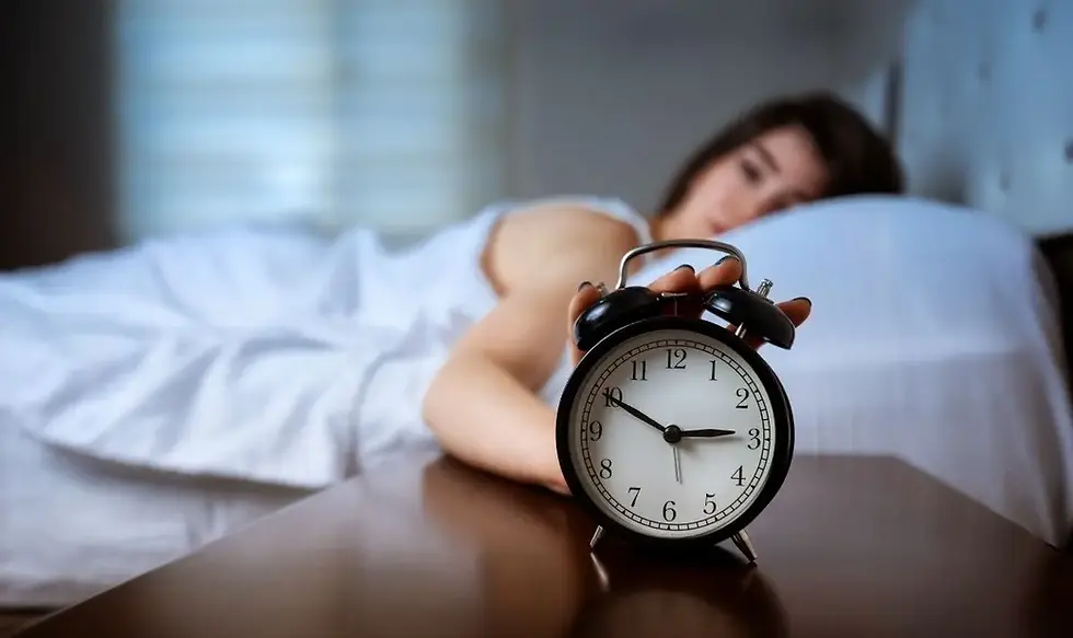 Vrouw ligt in bed en reikt naar een ouderwetse wekker op een houten tafel. Witte lakens, wazige achtergrond, ontwakende sfeer.