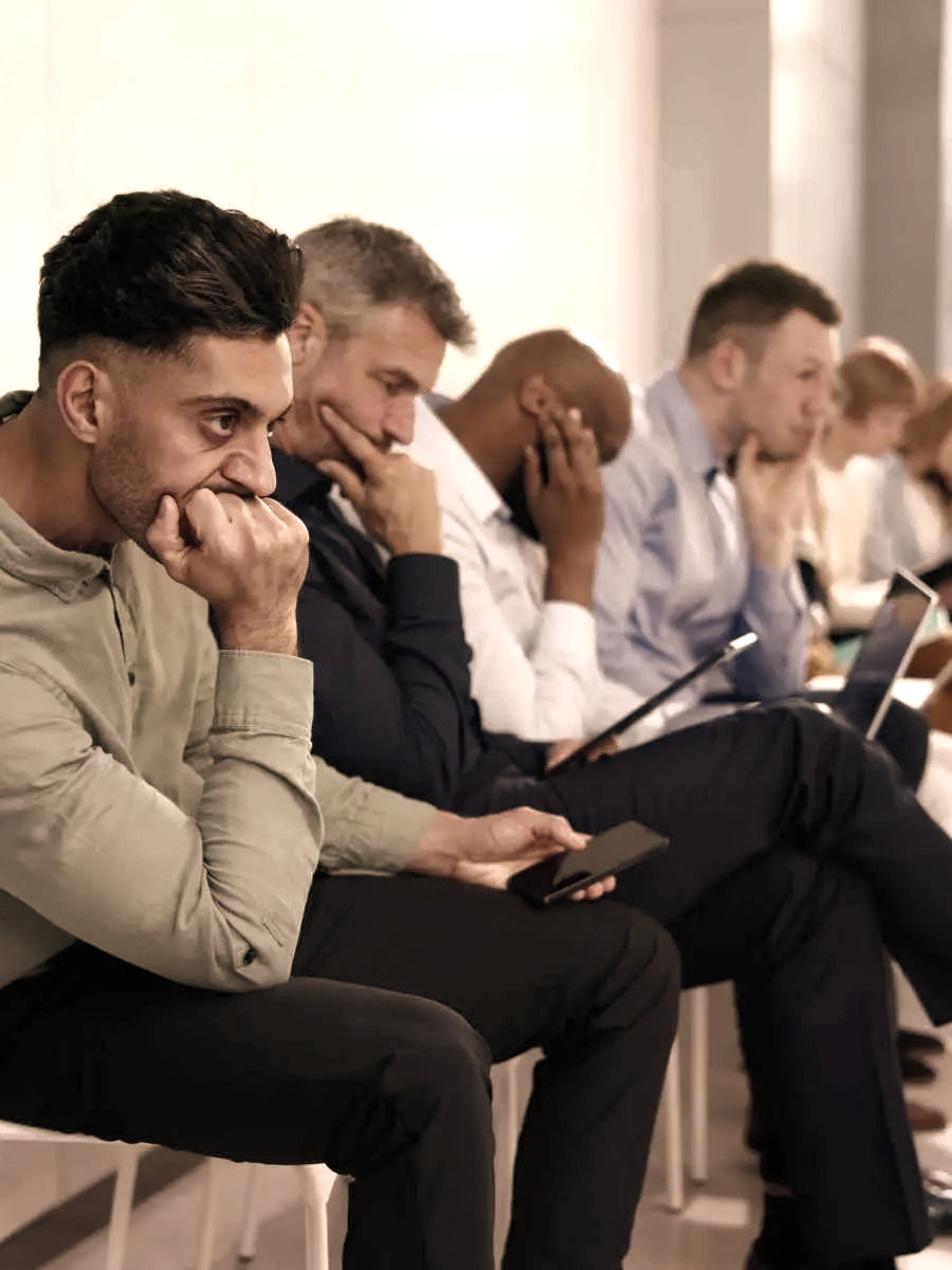 Mannen in formele kleding zitten op een rij en kijken bezorgd. De setting is een neutrale zaal. Velen houden hun kin of telefoon vast.