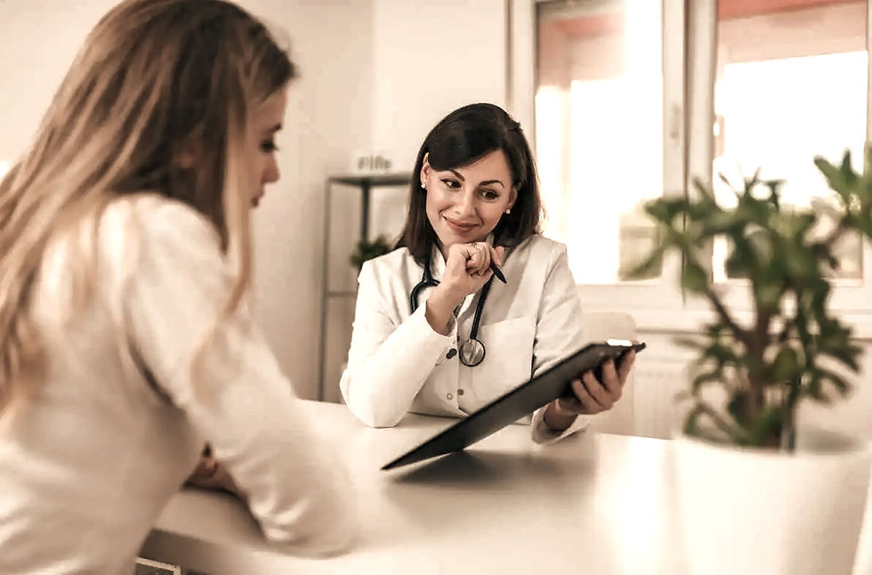 Twee vrouwen in gesprek aan een bureau in een lichte kamer. De vrouw rechts draagt een stethoscoop en houdt een schrijfblok vast, glimlachend.