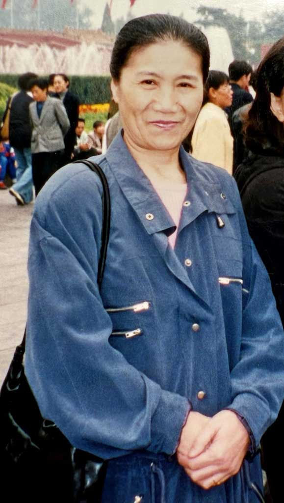 Vrouw glimlacht in een blauwe jas, staande op een drukke buitenlocatie met mensen op de achtergrond. Rustige sfeer.