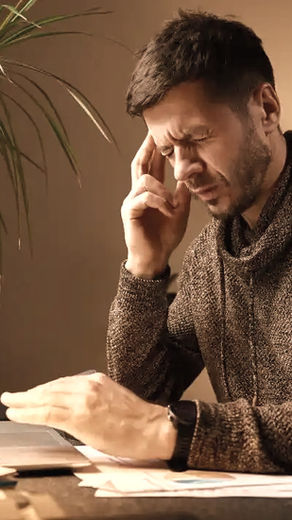 Man bij laptop, wrijft over zijn vermoeide hoofd. Plantenpot en bureaulamp op tafel. Rustige omgeving, warme kleuren.