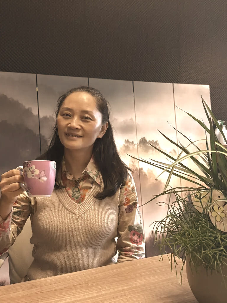 Vrouw glimlachend, houdt roze mok vast. Zittend bij houten tafel, plant en schilderij met bergachtergrond. Warme, rustige sfeer.