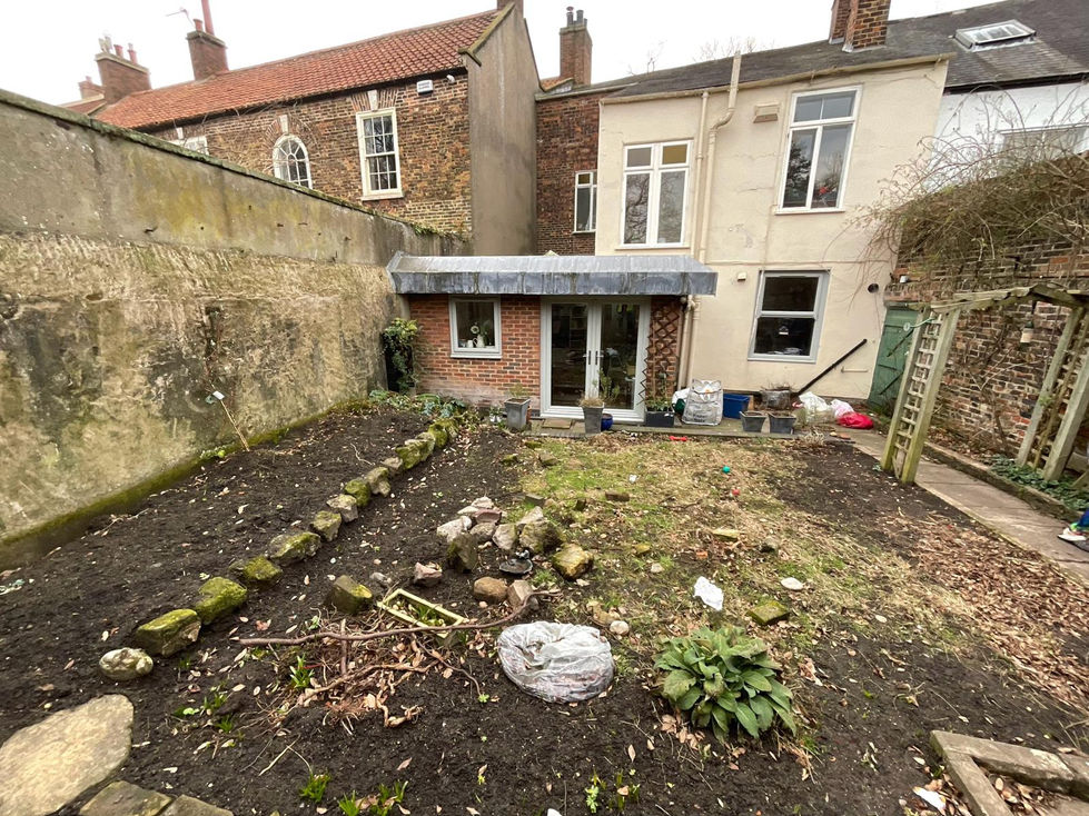 Overgrown townhouse garden with uneven ground, scattered debris, old planting areas and a worn pergola structure. Brick outbuilding on the right, tall boundary walls and a tired patio area leading to the rear of the house.