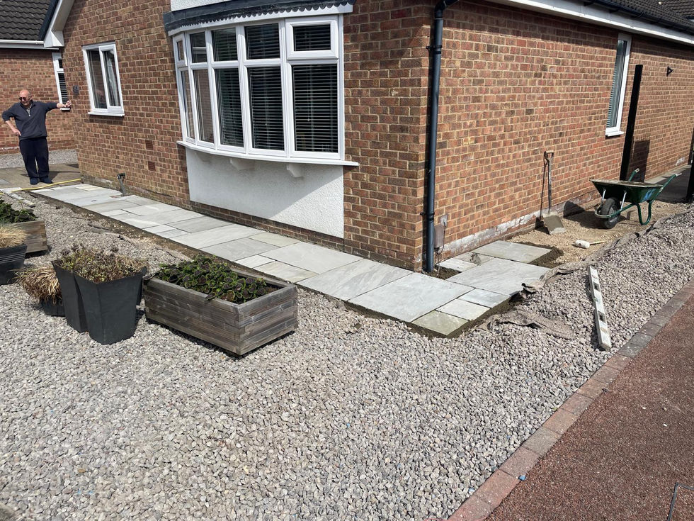 New sandstone paving being laid along the front of a red-brick house, surrounded by gravel and wooden planters with leafy green plants.