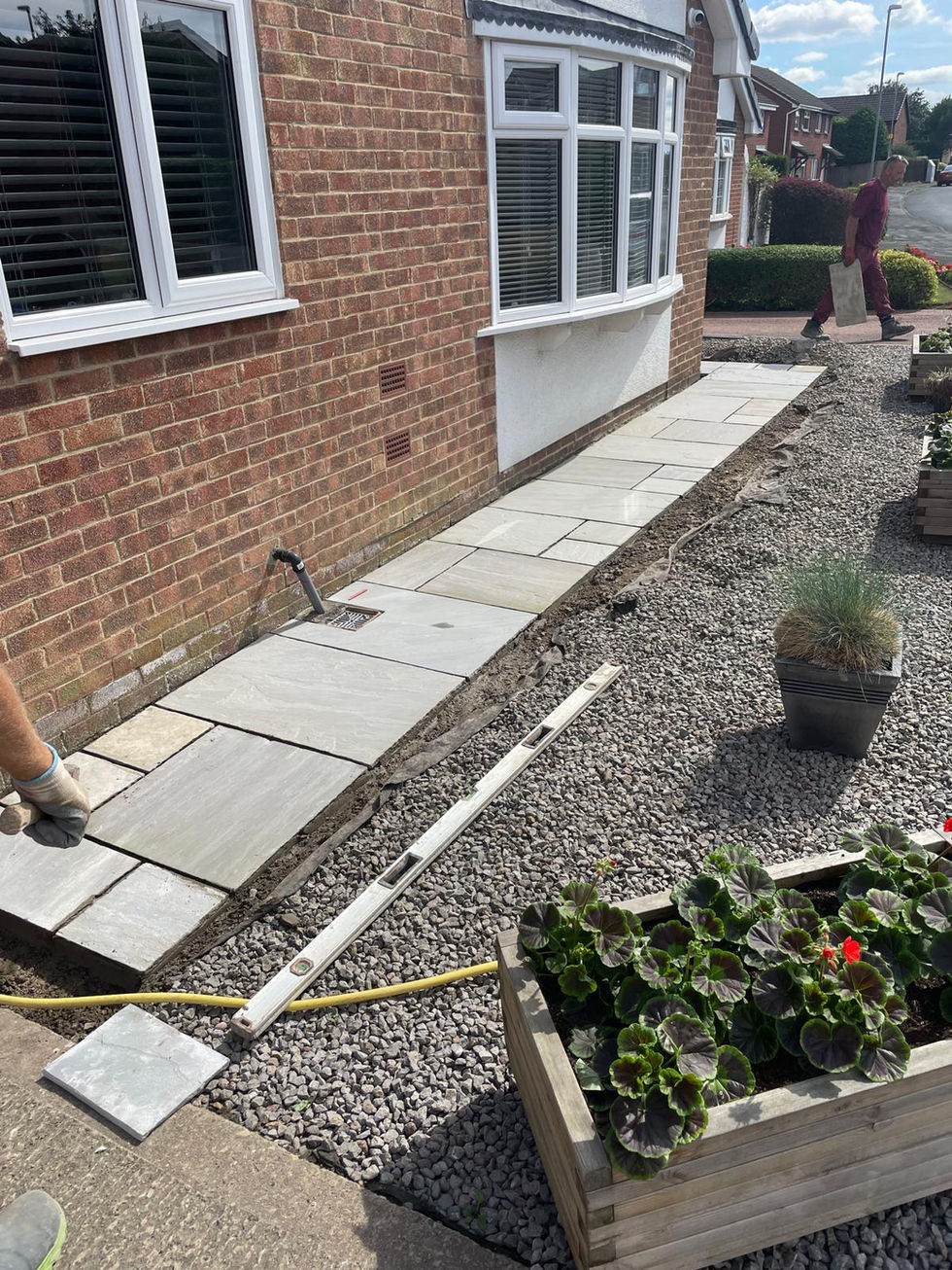 New sandstone paving being laid along the front of a red-brick house, surrounded by gravel and wooden planters with leafy green plants.