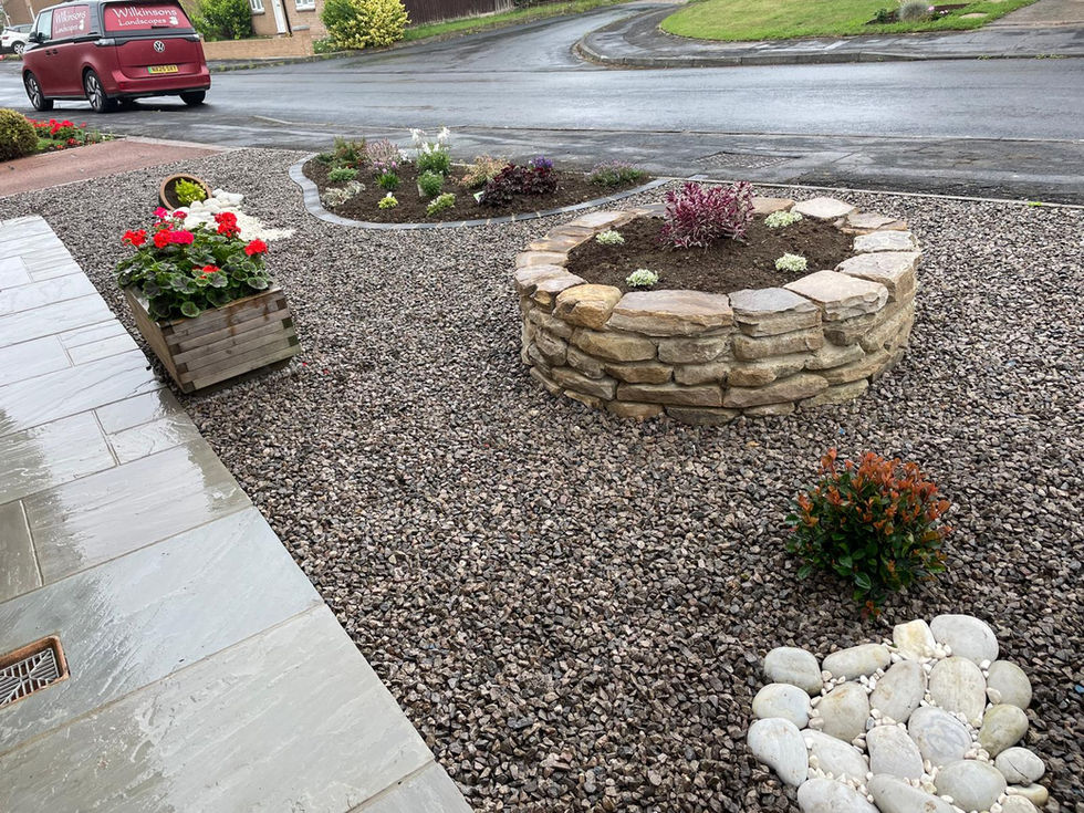 Front garden with circular raised stone planter, colourful flowers, and gravel beds outside a red-brick bungalow with white window frames.
