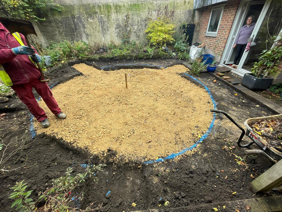 Circular patio area marked out with blue spray line and filled with compacted hardcore. Landscaping tools and workers preparing the site, with garden beds cleared around the edges.