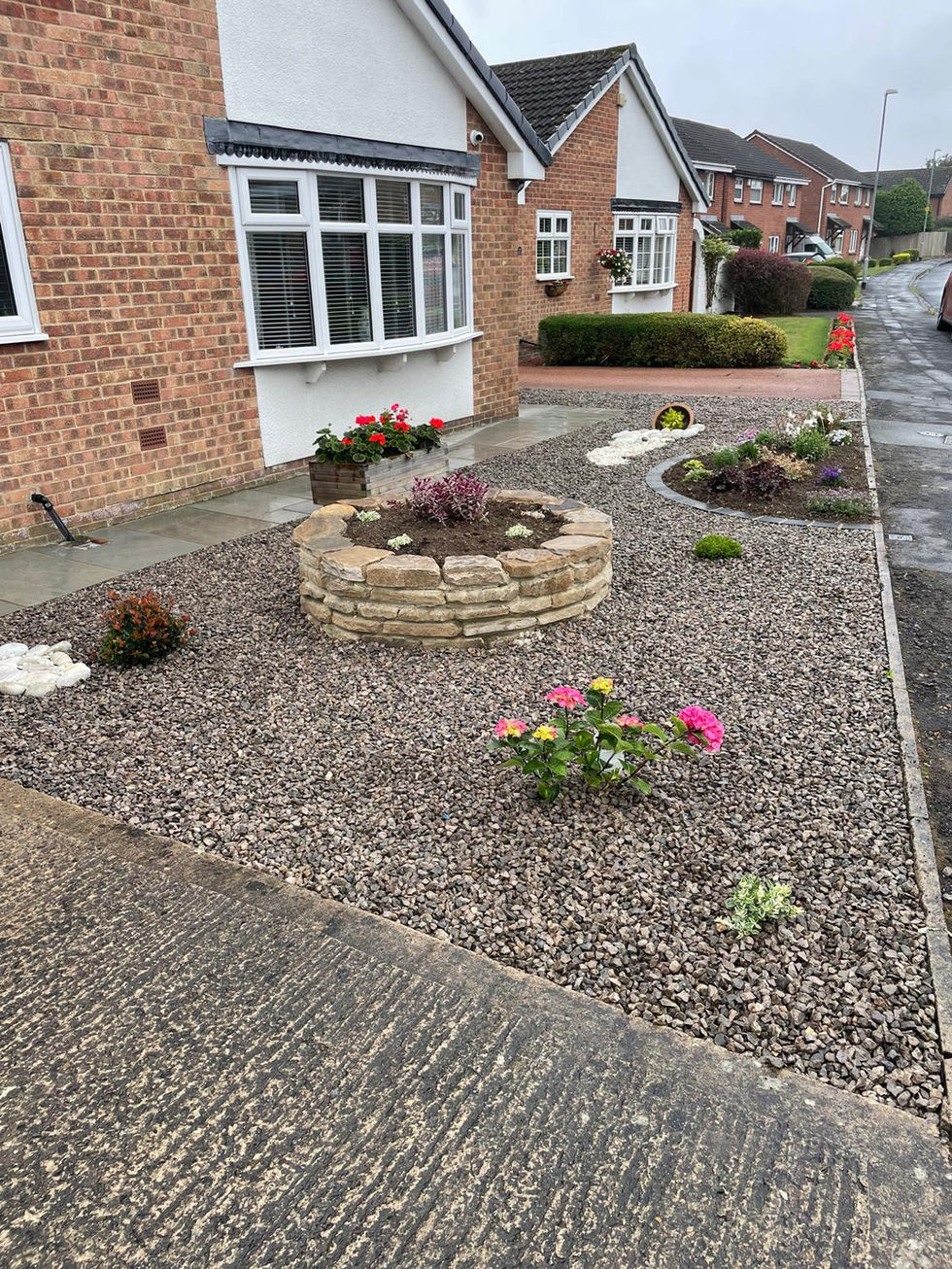 Front garden with circular raised stone planter, colourful flowers, and gravel beds outside a red-brick bungalow with white window frames.