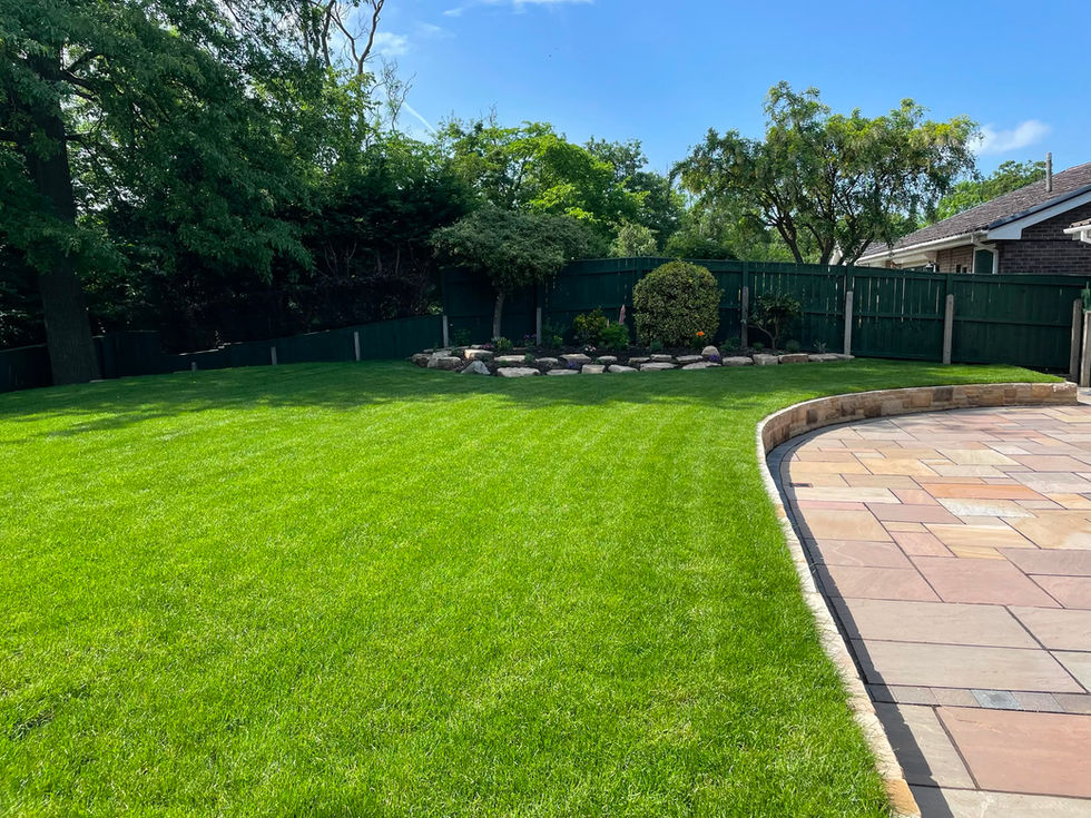 Newly established lawn and natural stone planting bed featuring seasonal flowers and specimen shrubs, set against mature trees and dark green fencing.