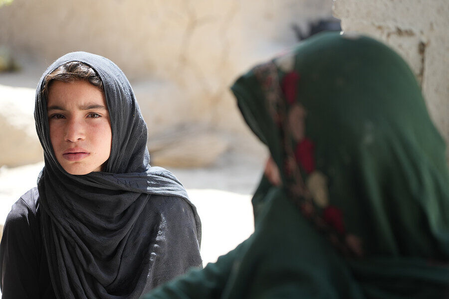 A girl wearing a gray headscarf sits facing the camera, with a woman in a green headscarf visible in the foreground.